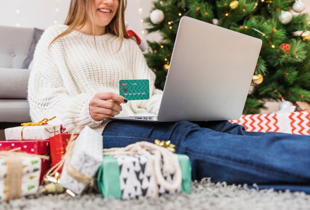A woman sitting by a Christmas tree shopping online with a laptop and credit card, surrounded by holiday gifts — Trendorashops Holiday Sale