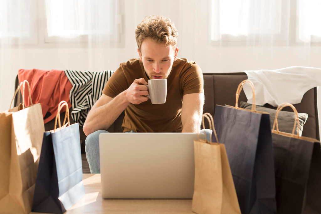 Man sitting at a table with a laptop and multiple shopping bags, drinking from a mug.