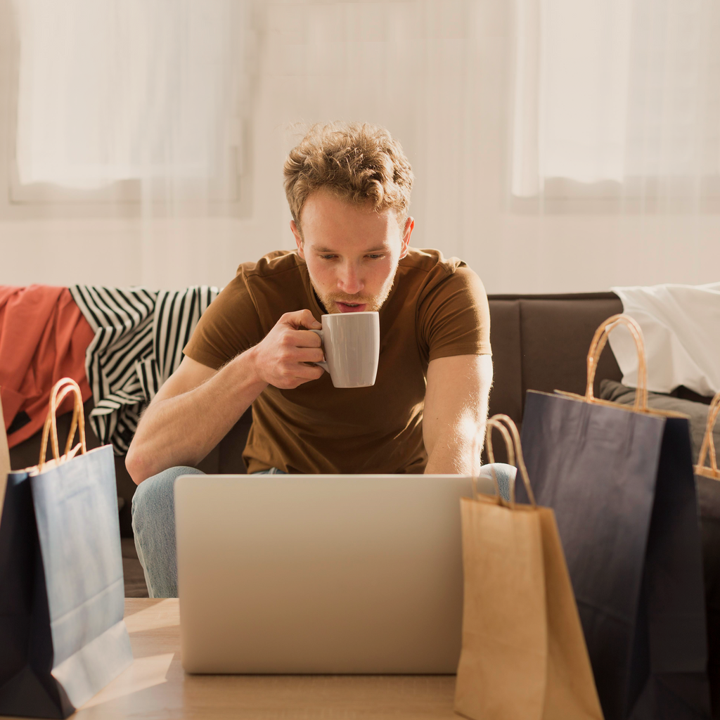 Man sitting at a table with a laptop and shopping bags, holding a mug.