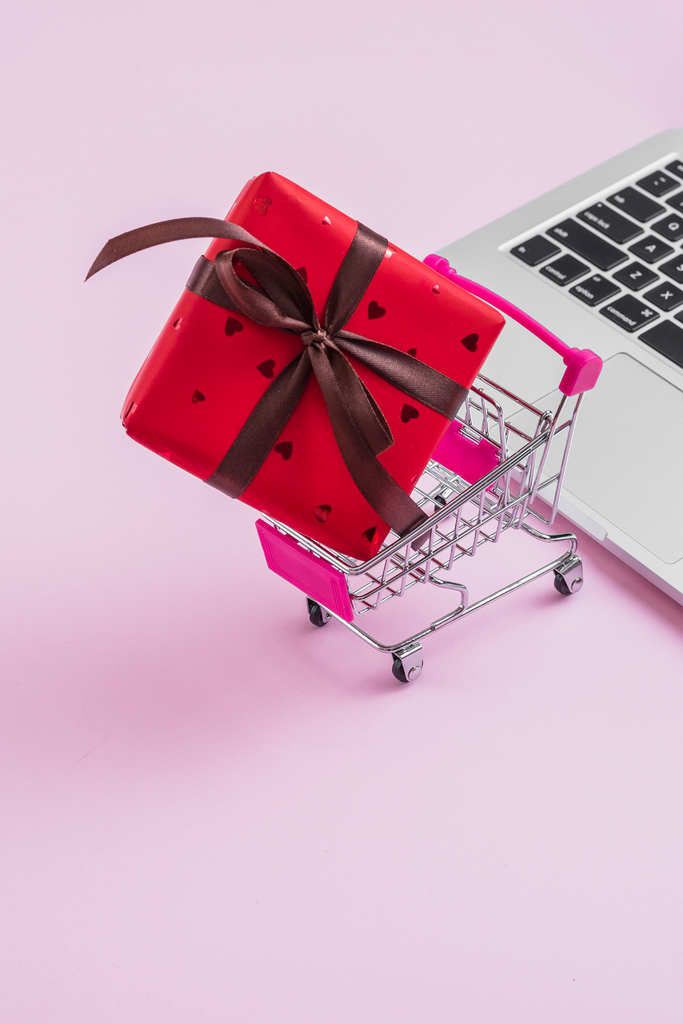 Red gift box with a brown ribbon in a small shopping cart next to a laptop on a pink background