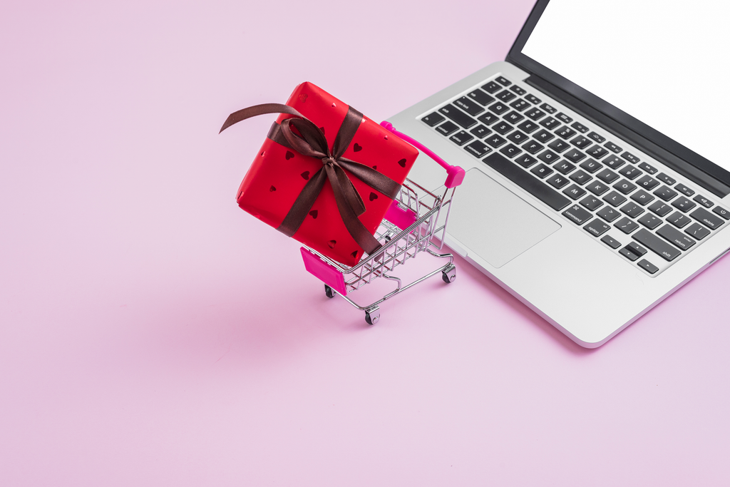 Red gift box with a brown ribbon in a small shopping cart next to a laptop on a pink background